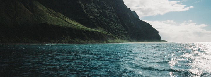 A vast body of water with sparkling sunlight on its surface stretches towards a distant, lush green mountain range under a partly cloudy sky.