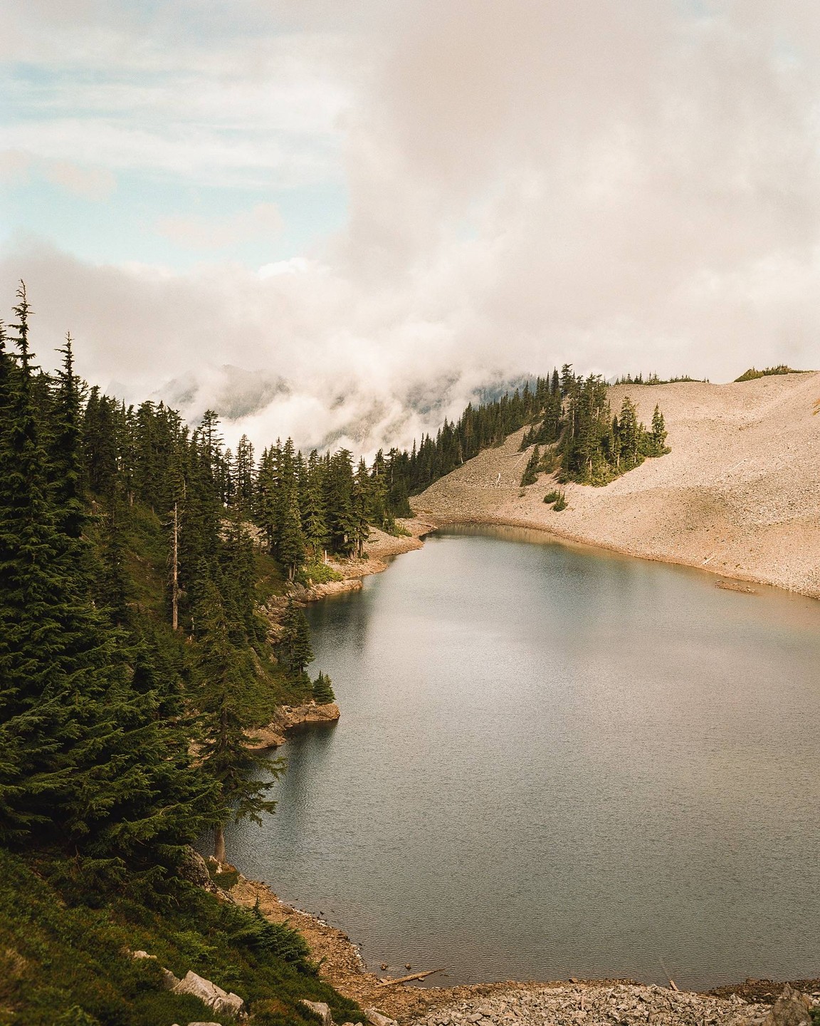 A lake is surrounded by a forest and a rocky hill, with fog covering the mountains in the background.