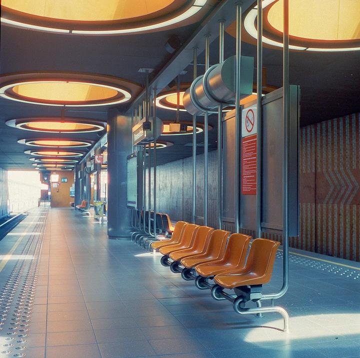 An empty subway platform with orange seats and circular overhead lights is illuminated by sunlight.