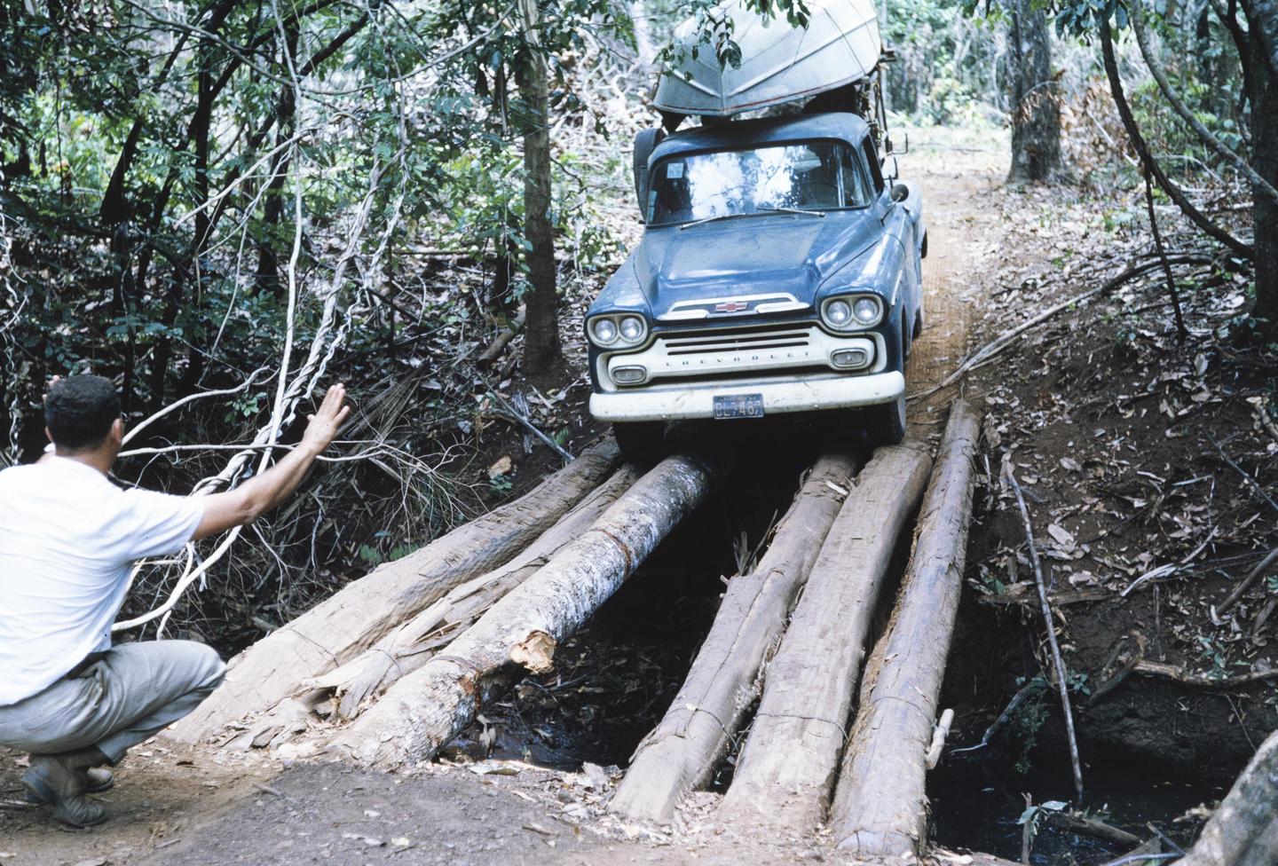 A man guides a blue truck with a boat on its roof over a log bridge in a forest.