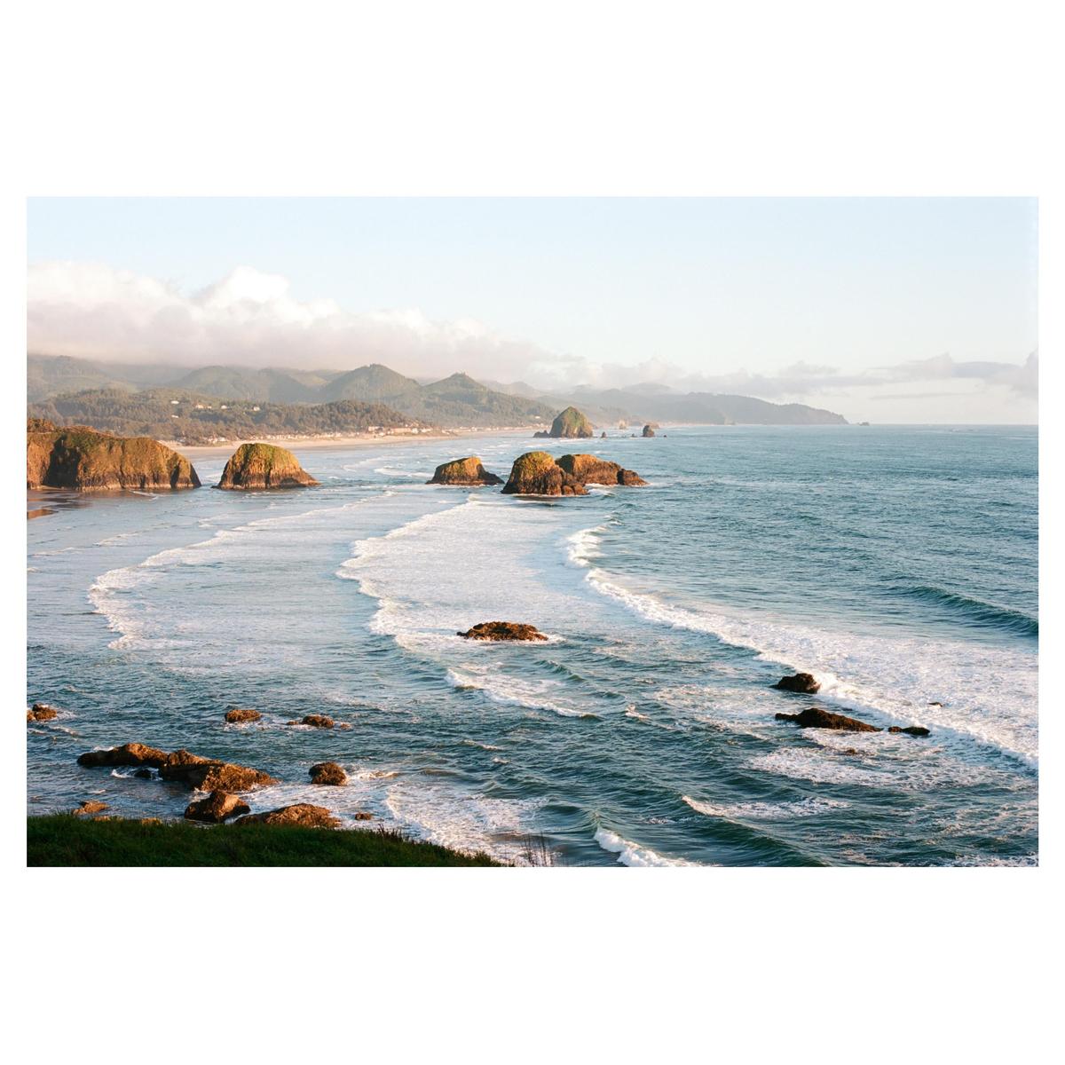 Waves crash on a rocky beach with hills and a town in the distance under a hazy sky.