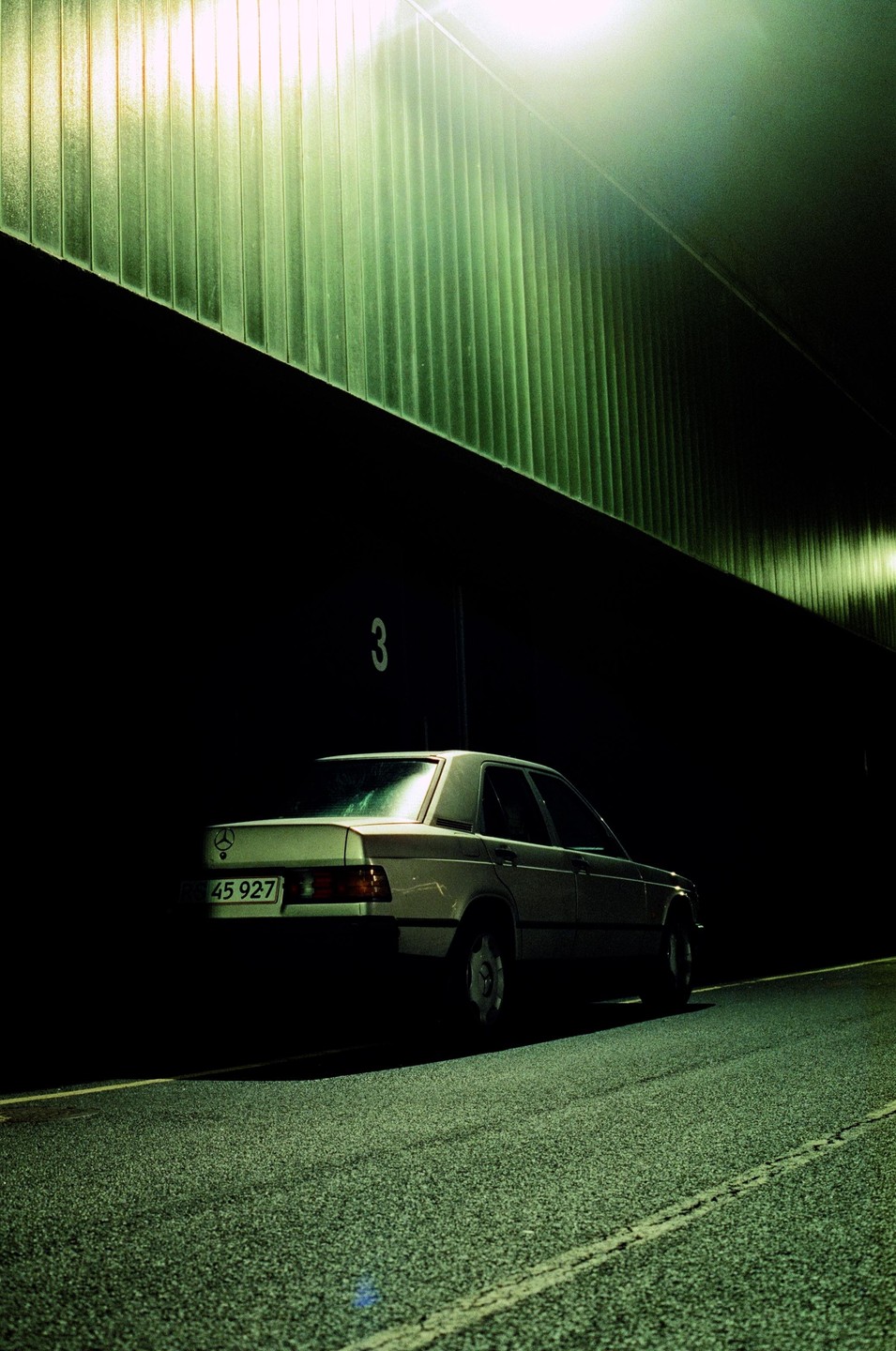 A car is parked on the side of a road next to a building with a green corrugated wall at night.