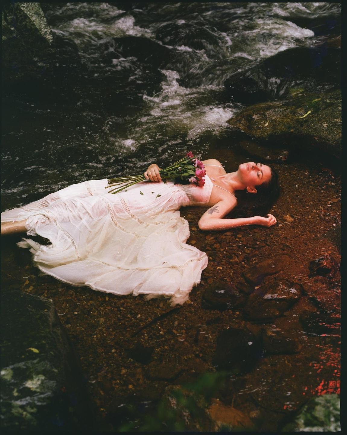 A woman in a white dress lies in a shallow stream with a bouquet of flowers on her chest.