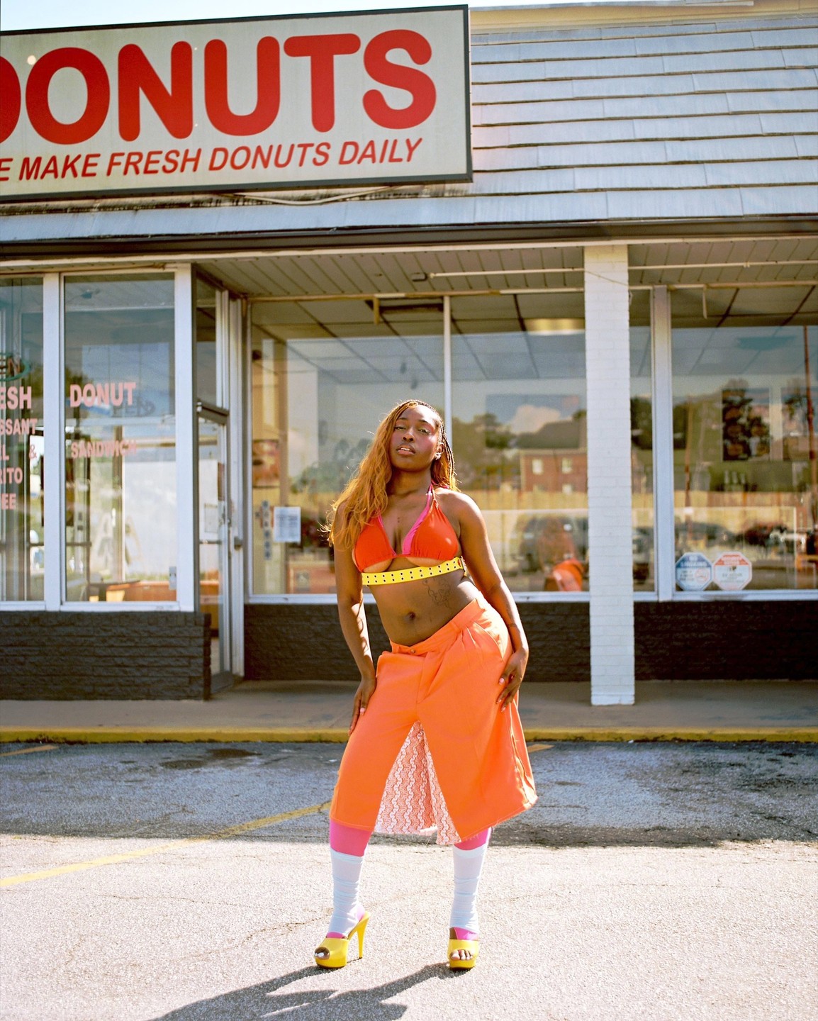 A woman in a bright orange and pink outfit stands in front of a donut shop.