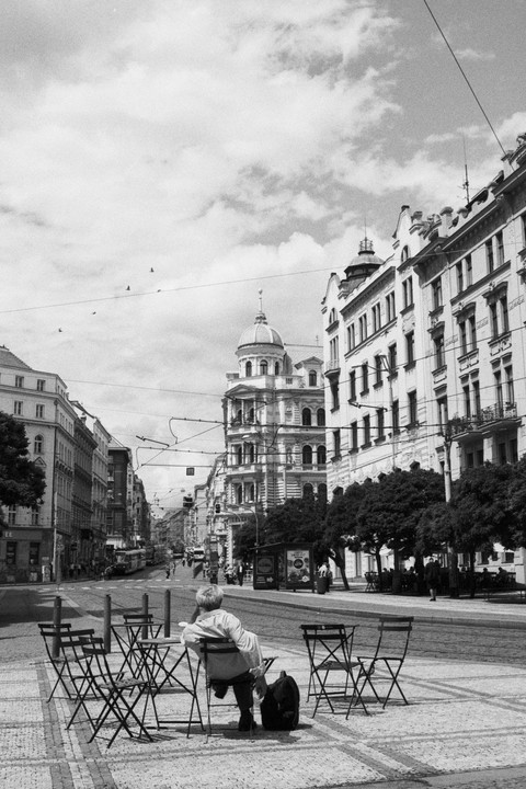 A person sits at an outdoor cafe table on a city street lined with ornate buildings and trees.