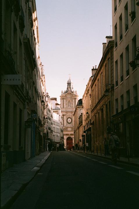 A narrow street in Paris leads to a church with a clock tower.