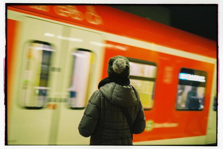 A person in a winter coat and hat stands on a platform next to a moving red and white train.