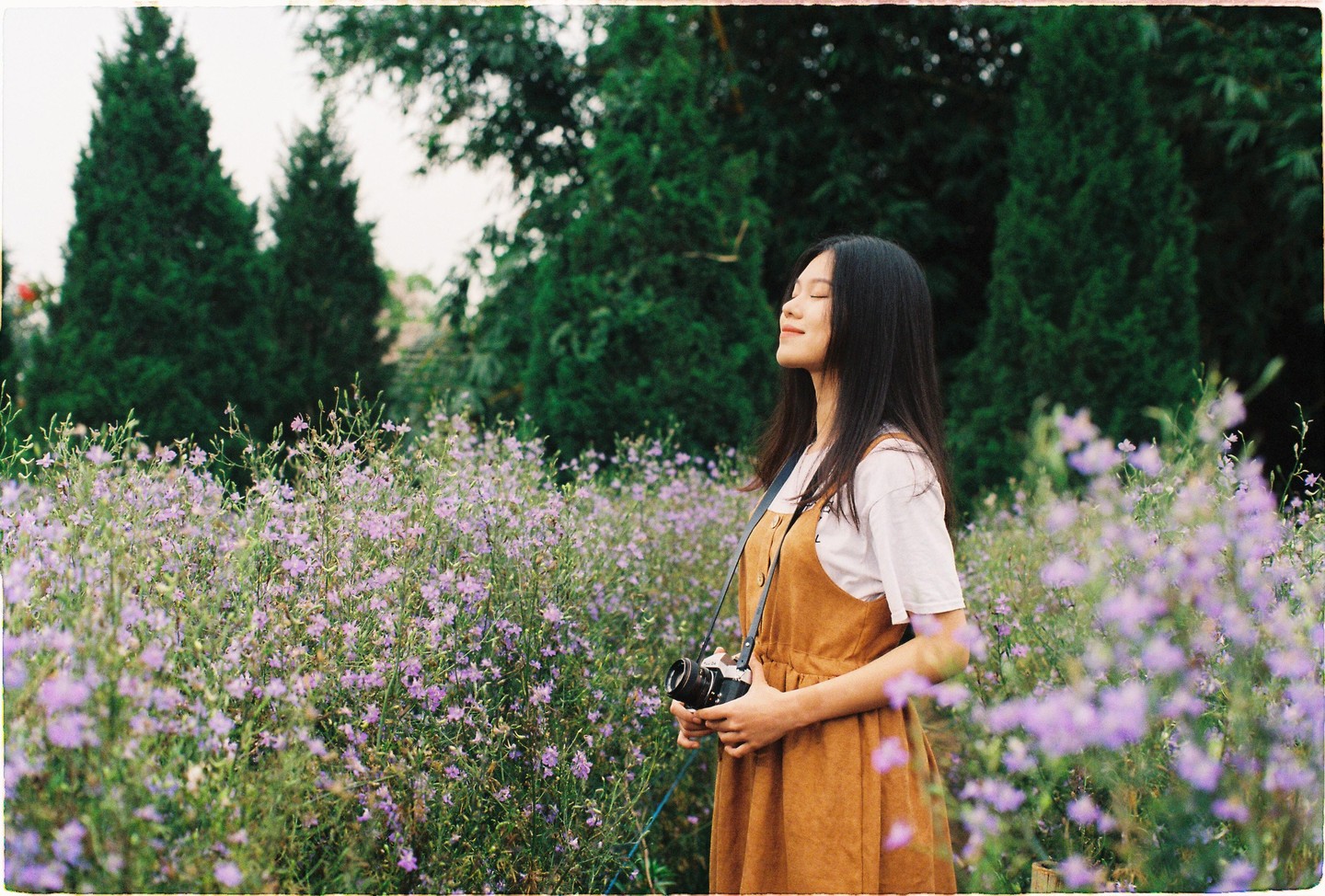 A woman stands in a field of purple flowers with her eyes closed and a camera in her hands.