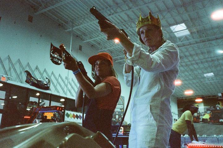 Two people play an arcade shooting game, one wearing a gold crown and the other an orange baseball cap.