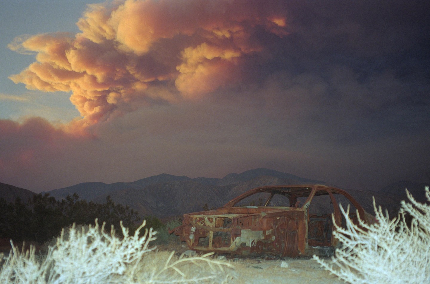 A rusty, burnt-out car sits in a desert landscape with mountains in the background and a large, orange smoke plume rising into the sky.