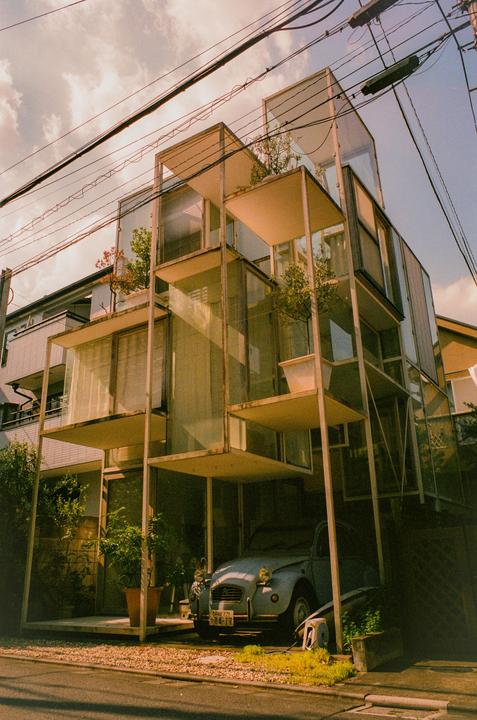 A light blue Citroen 2CV is parked under a modern glass and concrete building with plants on its balconies.
