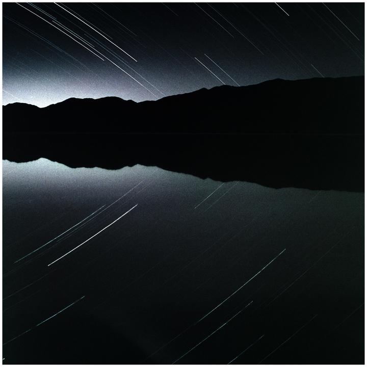 Star trails are visible above a mountain range reflected in a body of water at night.
