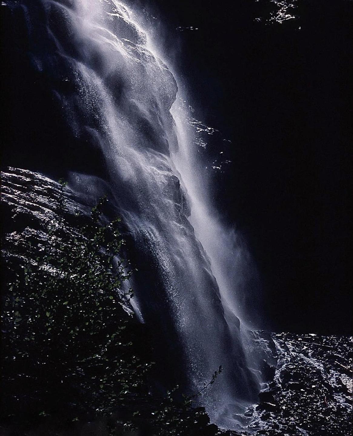 A waterfall cascades down a dark rock face with some foliage visible in the foreground.