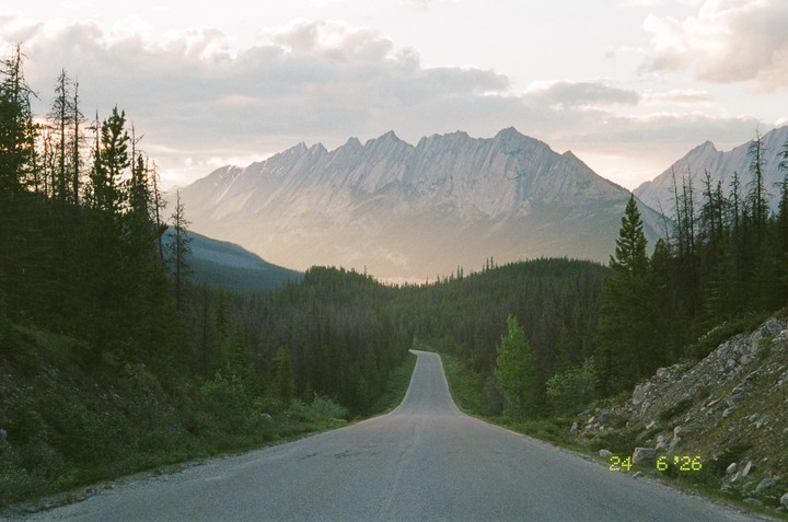A winding road leads through a forest towards mountains under a cloudy sky.