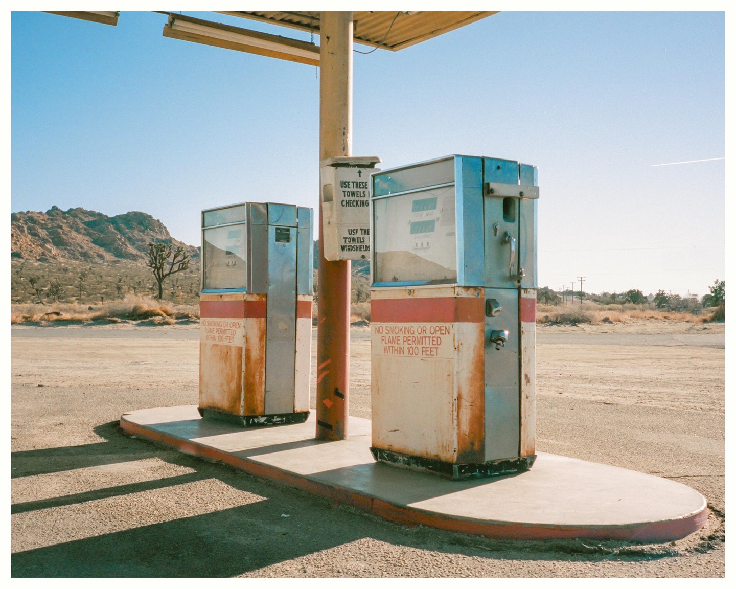 Two old gas pumps stand on a concrete island in a desert landscape with mountains in the background.