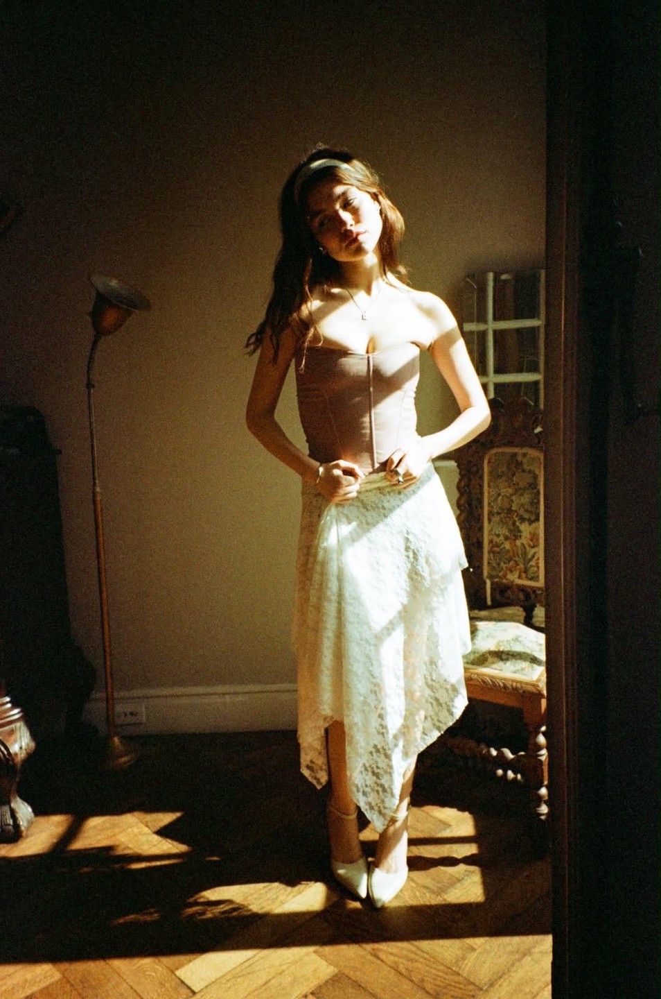 A woman stands in a sunlit room, wearing a corset and a white lace skirt, with a floor lamp and an ornate chair visible.