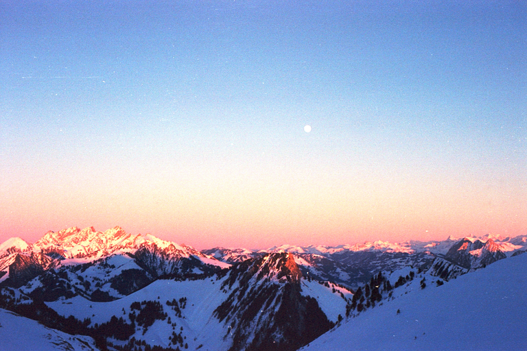 A full moon rises over snow-covered mountains with pink-hued peaks under a gradient sky.