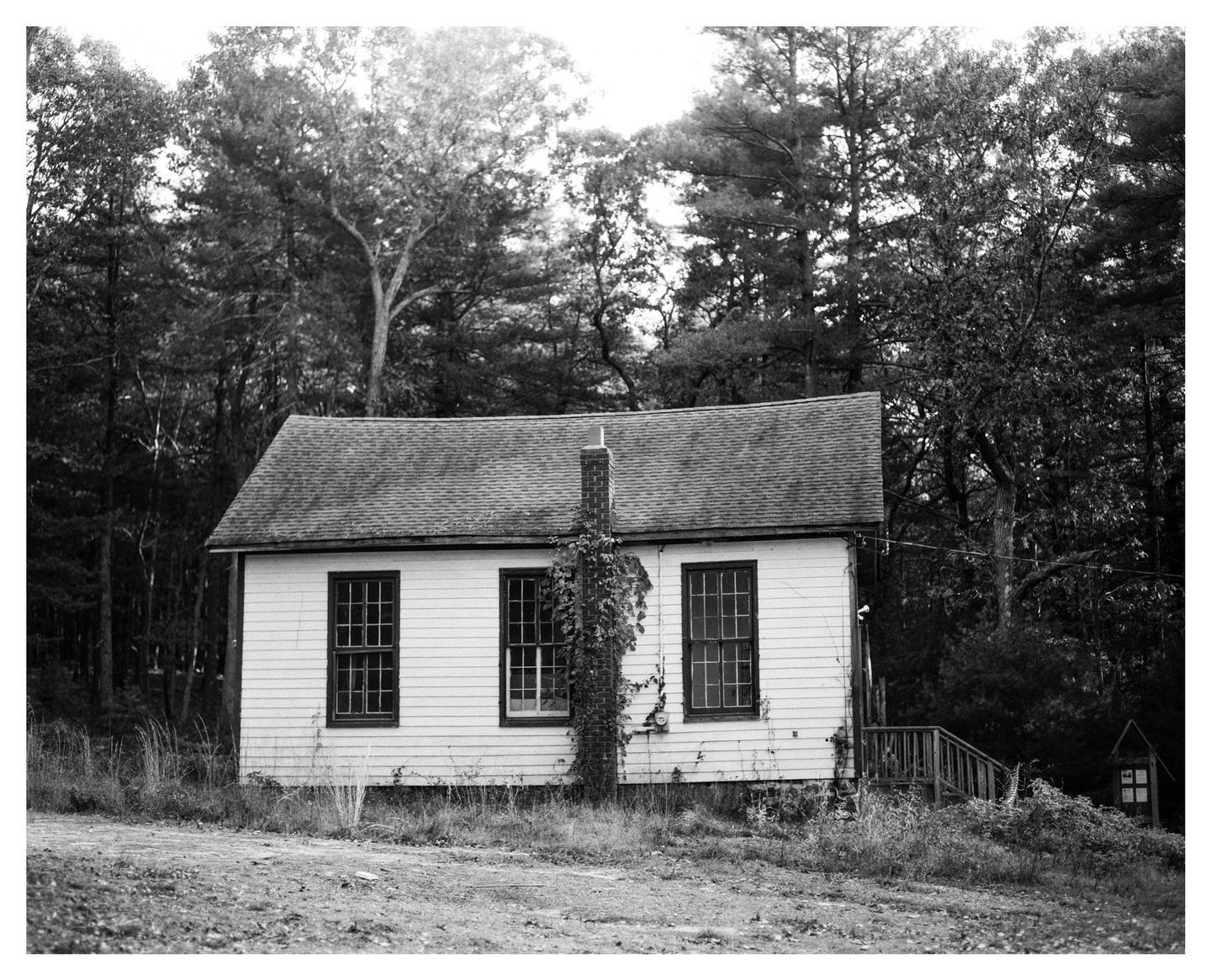 An abandoned house with a chimney and three windows stands in a clearing surrounded by trees.
