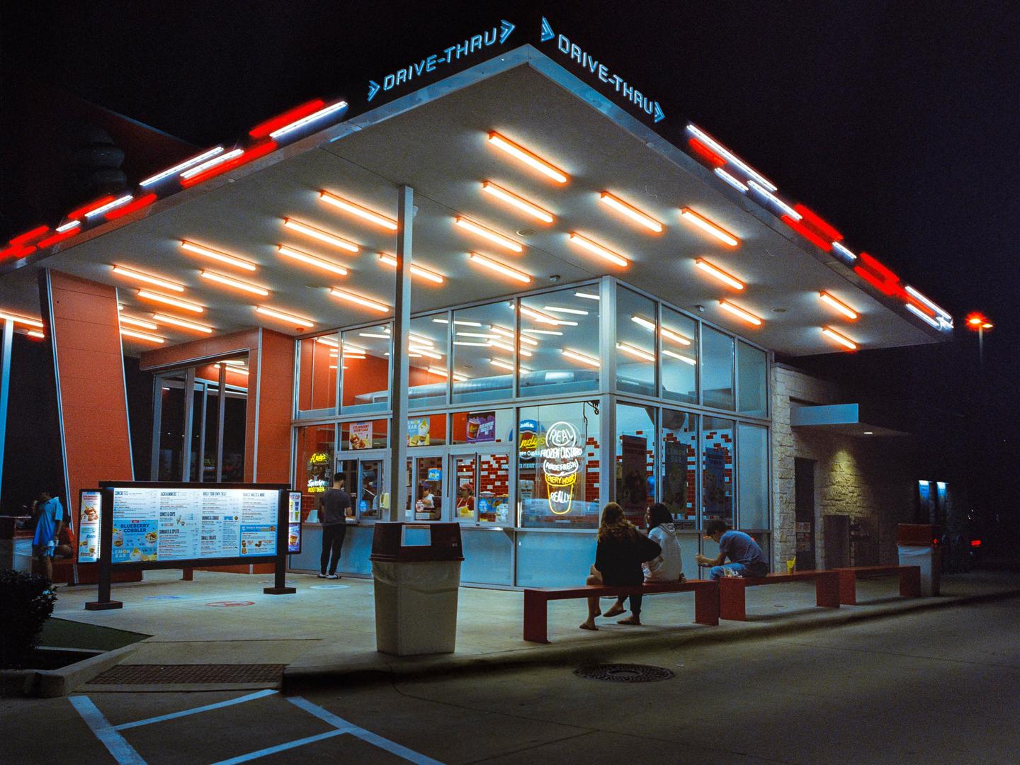 A group of people are gathered outside a brightly lit drive-thru restaurant at night.