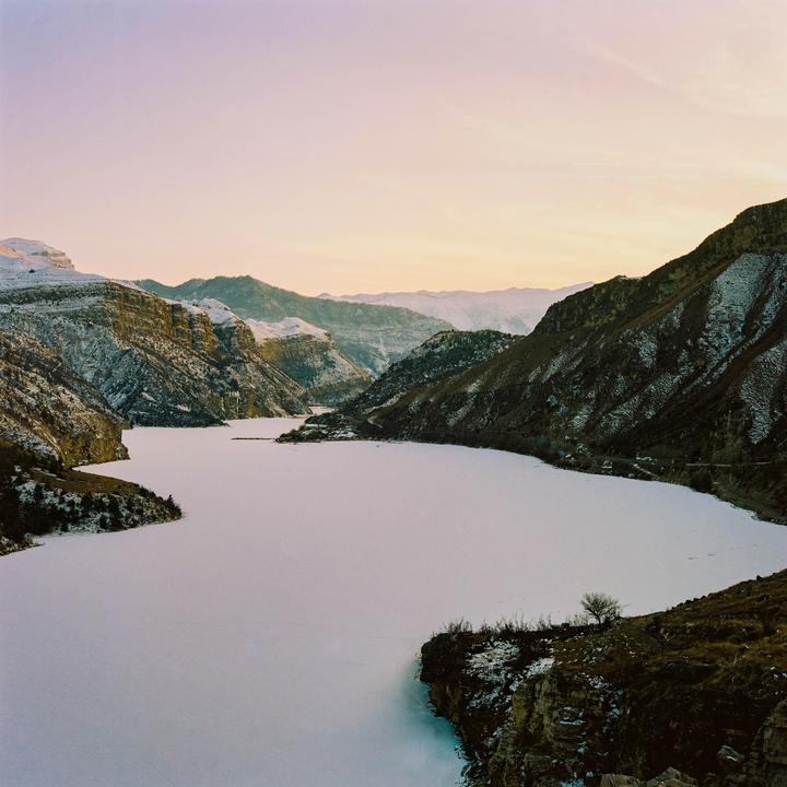 A frozen lake is surrounded by snow-dusted mountains under a pale sky at dusk.