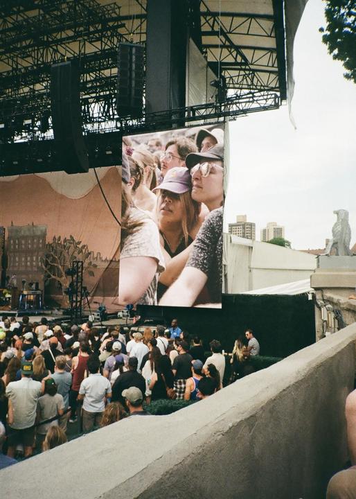A large crowd of people watches a concert with a stage and a large screen displaying audience members.