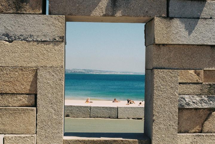 A beach with people sunbathing is visible through a rectangular opening in a stone wall.