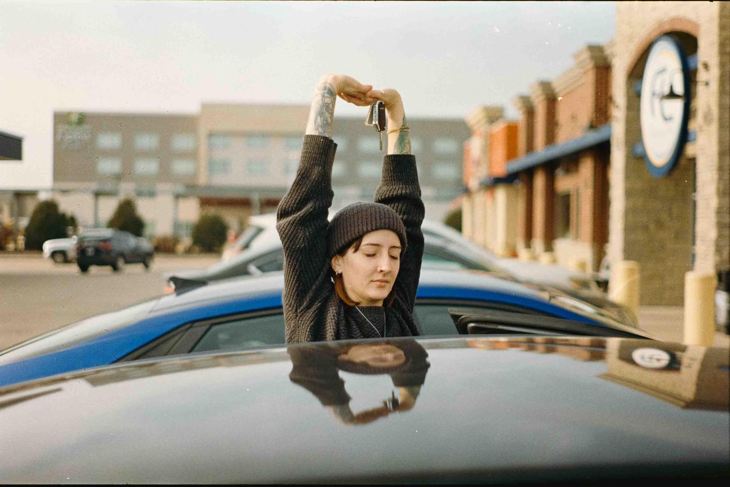 A woman with her eyes closed and arms raised above her head, holding keys, stands in a car's sunroof with her reflection visible on the car's roof.