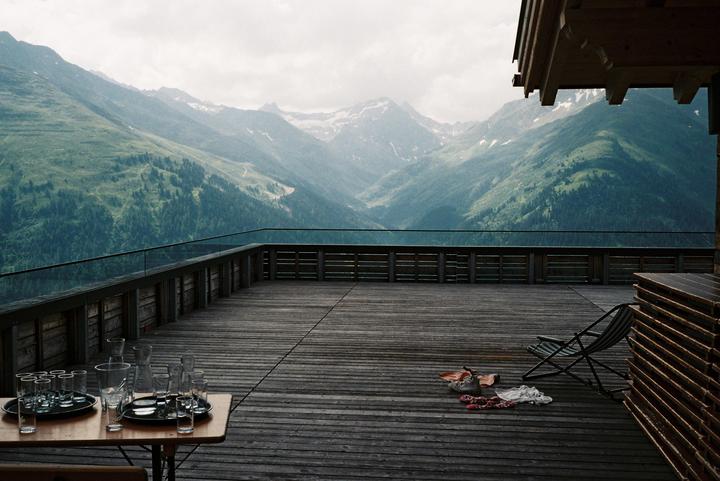 A wooden deck with a table of glasses and a lounge chair overlooks a mountain range.