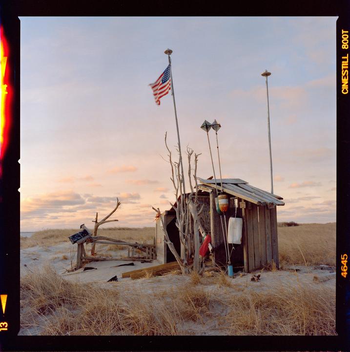 An American flag flies above a small wooden shack on a sandy beach with dry grass under a pastel sky.