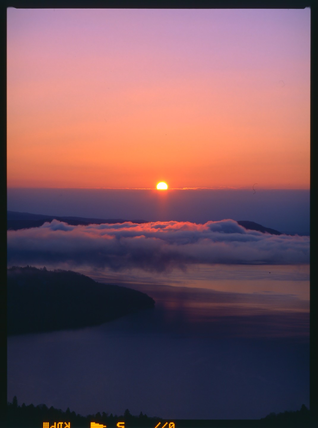 The sun rises over a lake with clouds reflecting the orange light, and a dark landmass is visible in the foreground.