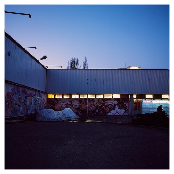 A building with illuminated windows and murals on its facade stands under a twilight sky, with a pile of snow in the foreground.