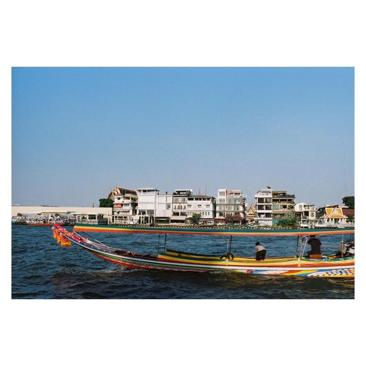 A colorful longtail boat with two men on board travels on a river with buildings on the far bank.