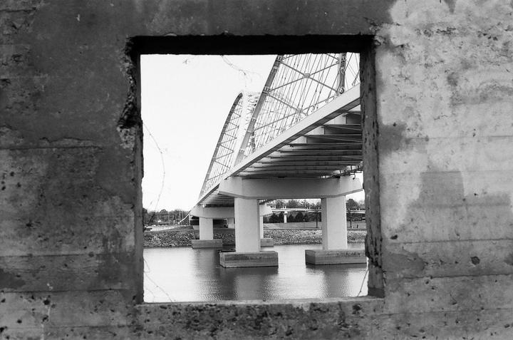 A bridge with a distinctive arch design spans a body of water, viewed through a rectangular opening in a concrete wall.