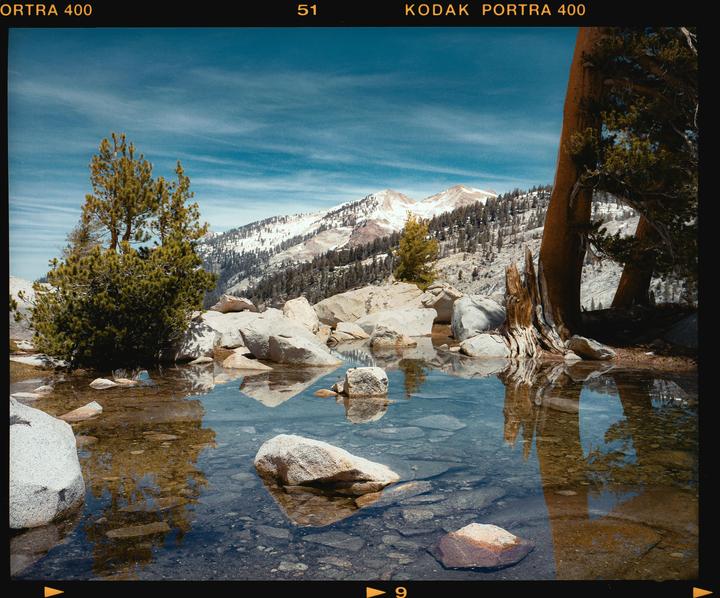 A clear mountain lake reflects trees and a snowy mountain under a blue sky.
