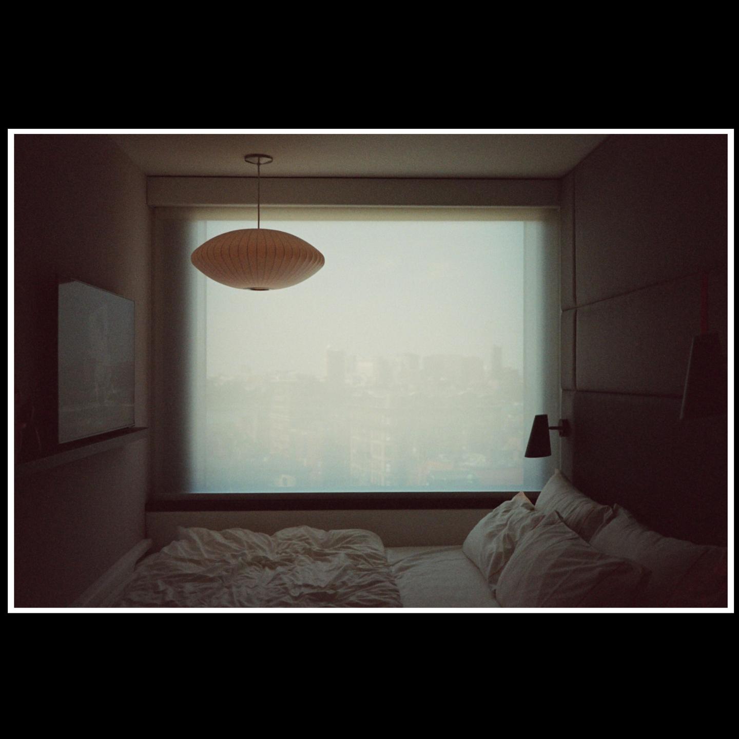 A bedroom with an unmade bed and a large window overlooking a city skyline.