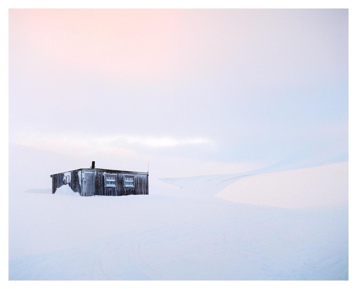 A small wooden cabin is surrounded by snow under a pale sky.