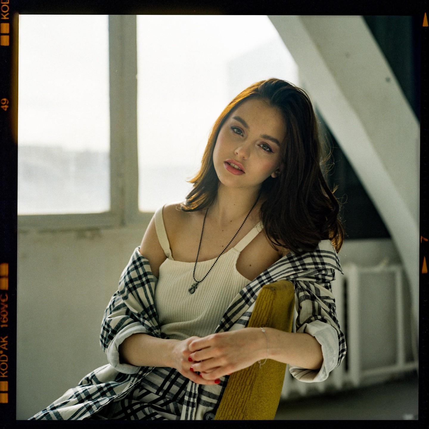 A woman with long dark hair and a plaid shirt sits on a yellow chair in front of a window.