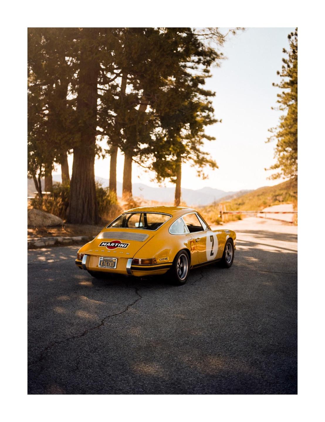 A yellow vintage sports car is parked on an asphalt road surrounded by tall trees and mountains in the distance.