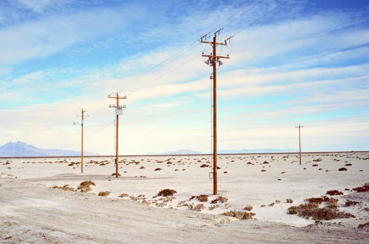 Several utility poles with power lines extend across a vast, flat desert landscape under a partly cloudy sky.