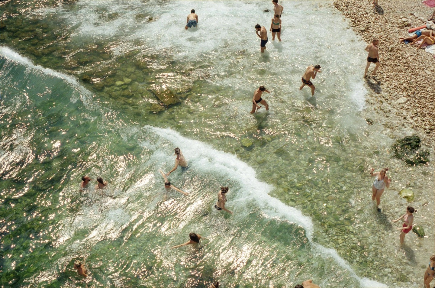 A group of people are swimming and wading in clear, shallow water next to a rocky beach.