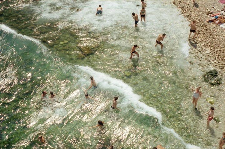 A group of people are swimming and wading in clear, shallow water next to a rocky beach.