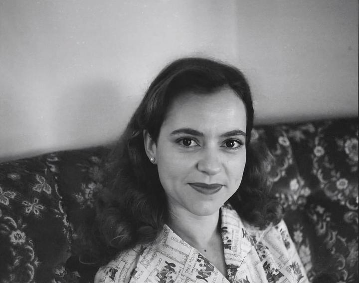 A woman with dark hair and red lipstick looks directly at the viewer while sitting on a patterned sofa.