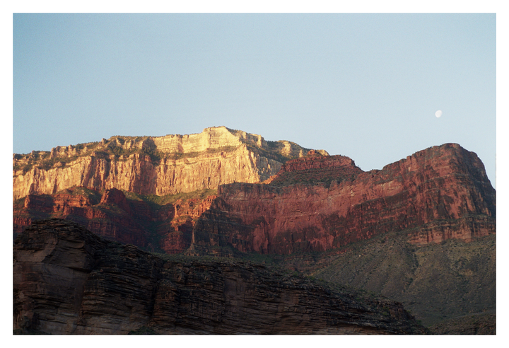 The Grand Canyon is illuminated by the morning sun, with the moon visible in the sky.