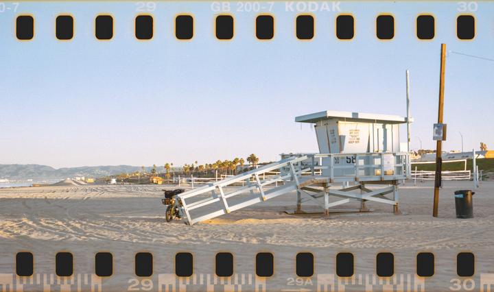 A lifeguard tower stands on a sandy beach with palm trees and mountains in the distance.