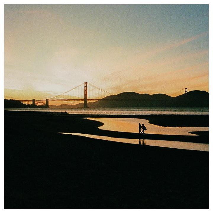 Two people stand in a tidal pool on a beach with the Golden Gate Bridge in the background at sunset.