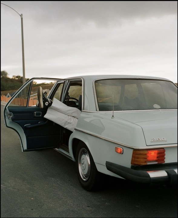 A person's legs and feet in sneakers are sticking out of the open door of a light blue vintage car parked on the side of a road.