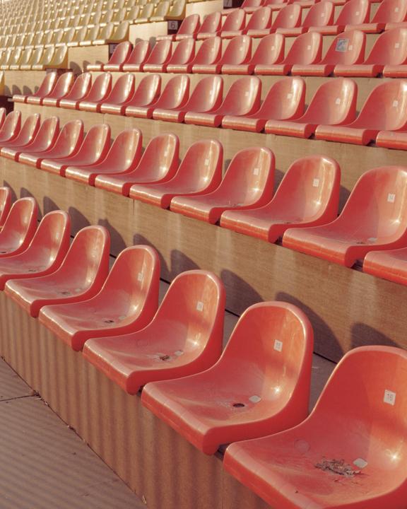 Rows of empty orange and yellow stadium seats are illuminated by sunlight.
