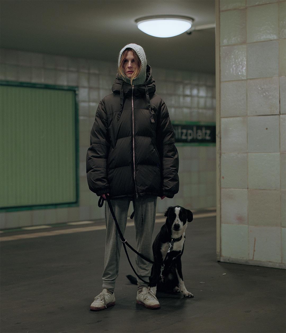 A woman in a puffer jacket and a dog on a leash stand in a subway station.