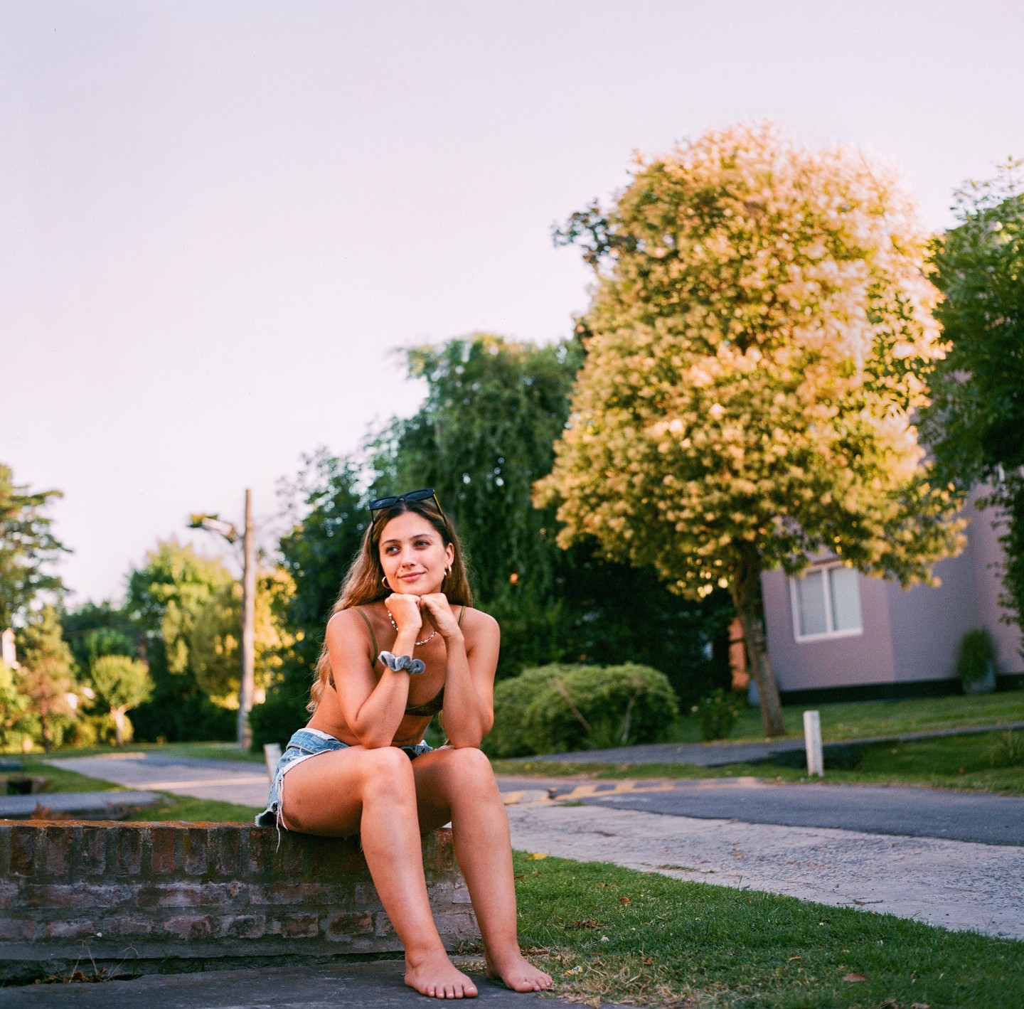 A woman sits barefoot on a brick wall, looking to the side with trees and a house in the background.