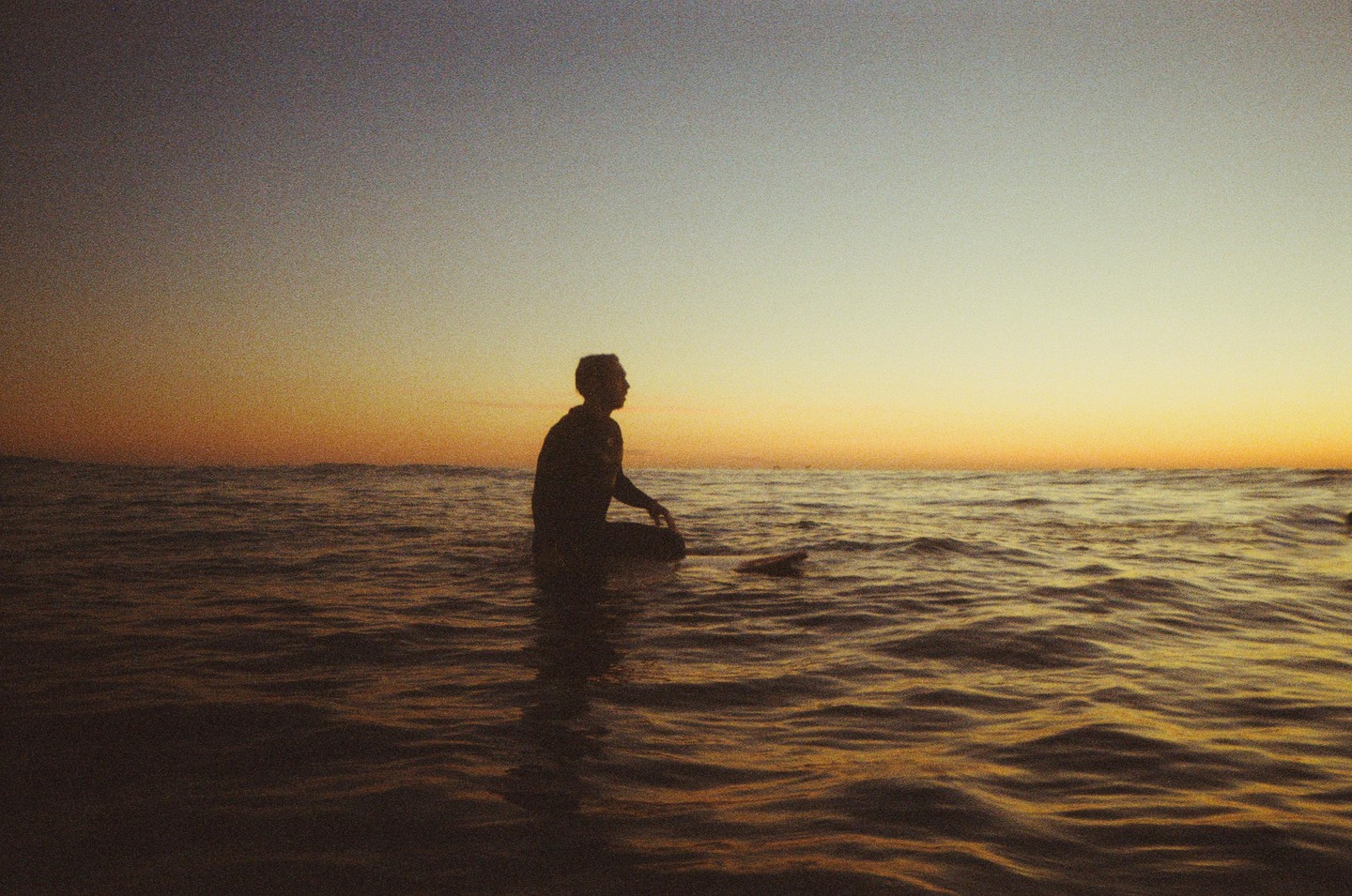 A man sits on a surfboard in the ocean at sunset.